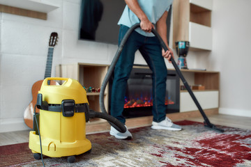 Wall Mural - Cropped view of young man cleaning the carpet with vacuum cleaner while working in the modern living room. Professional cleaner at work