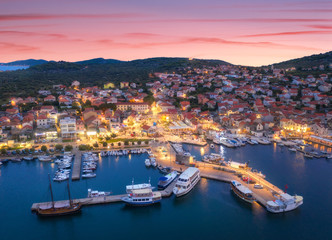 Wall Mural - Aerial view of boats and yachts in port and city at night. Summer landscape with city lights, buildings, illuminated streets, mountain, motorboats, blue sea, colorful sky at sunset. Top view. Croatia
