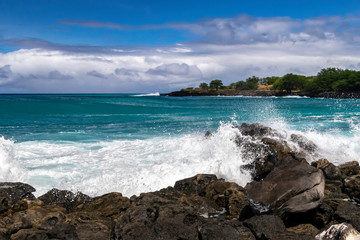 Wall Mural - Wave breaking on rough shore of the Kona coast, on Hawaii's Big Island. Blue-green Pacific ocean beyond; rocky shoreline with trees in the distance. Cloudy blue sky above.