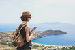 © kite_rin - Young tourist woman using smartphone over beautiful landscape with sea in Greece. Traveler girl with backpack and mobile phone. Travel, tourism, summer holidays, active lifestyle concept