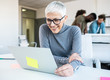 © Sanja - A senior employee looking at a lap top in an office with a few of her colleagues standing behind her, talking.