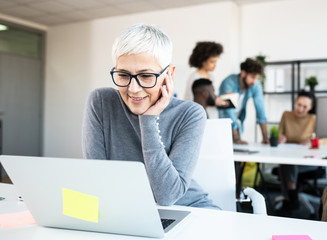 A senior female using her computer in an office. There are a few employees behind her at another table.