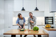 © Dusan Petkovic - Young charming smiling caucasian woman in apron standing in kitchen and cutting cucumber while talking with her boyfriend. Man holding cherry tomato and talking about healthy lifestyle.