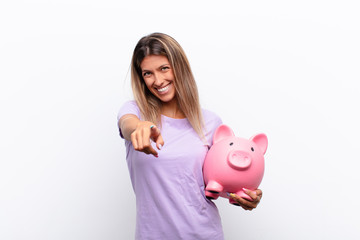 young pretty woman pointing at camera with a satisfied, confident, friendly smile, choosing you with a piggy bank.