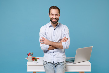 Smiling young bearded man in light shirt work near white desk with pc laptop isolated on pastel blue background. Achievement business career lifestyle concept. Mock up copy space. Hold hands crossed.