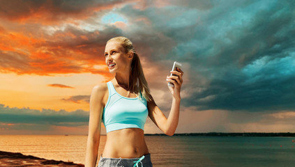 Wall Mural - fitness, sport, technology and people concept - smiling young woman with smartphone exercising over sea and sunset sky on background