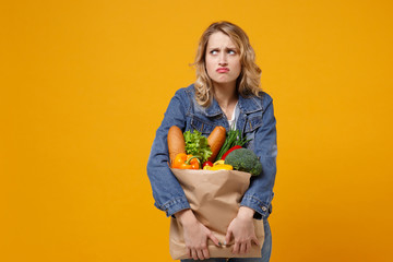 Wall Mural - Displeased young woman in denim clothes isolated on yellow orange background. Delivery service from shop or restaurant concept. Holding brown craft paper bag for takeaway mock up with food products.