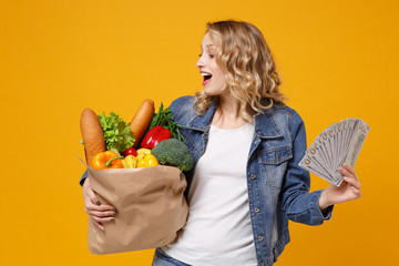 Wall Mural - Amazed young girl in denim clothes isolated on orange background. Delivery service from shop or restaurant concept. Hold brown craft paper bag for takeaway mock up with food product fan of cash money.