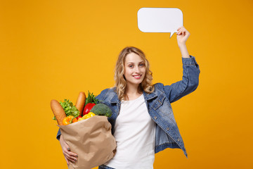 Wall Mural - Smiling girl in denim clothes isolated on orange background. Delivery service from shop or restaurant concept. Hold brown craft paper bag for takeaway mock up with food products empty blank Say cloud.