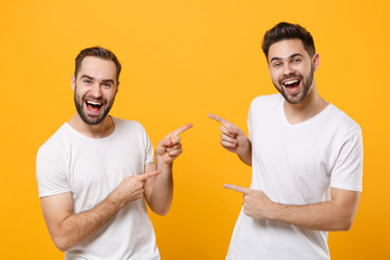 Wall Mural - Cheerful young men guys friends in white blank empty t-shirts posing isolated on yellow orange background in studio. People lifestyle concept. Mock up copy space. Pointing index fingers at each other.