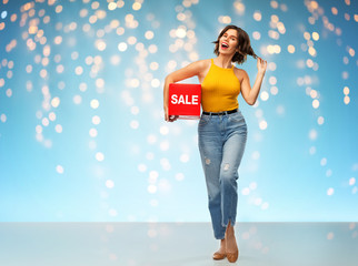 Wall Mural - shopping and people concept - happy smiling young woman in mustard yellow top and jeans with sale sign posing over holidays lights on blue background