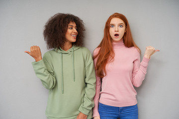 Wall Mural - Indoor shot of attractive young women rasing their hands and pointing in different directions, showing various emotions while standing over grey background