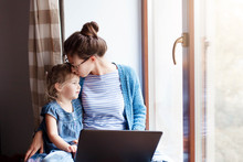 Mother Hug And Kiss Daughter. Working Mom Works From Home Office. Woman And Cute Child Sitting On Window Sill. Freelancer Using Laptop And The Internet. Female Business, Kindness, Love. Copy Space