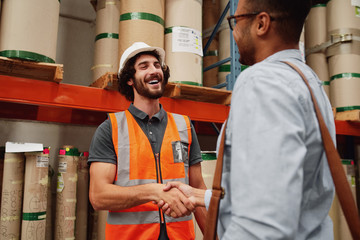 Smiling warehouse manager in orange jacket uniform and helmet greeting business partner and shaking hands