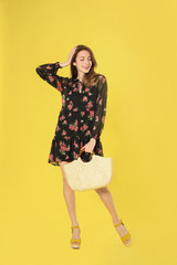 Canvas Print - Young woman wearing floral print dress with straw bag on yellow background