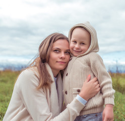 Young family, woman mother happy smiling, little boy sweater hood laughing rejoices, rest field, parenting child care love, parental support. Autumn spring day. Warm clothing.