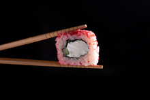 Chopsticks Holding Seaweed Pink Roll Filled With Avocado And Fish Over Black Background