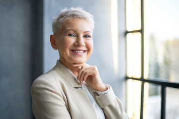 Wall Mural - Happy middle aged blonde woman in stylish beige jacket standing by window in office, holding hand under her chin and smiling at camera. Success, business, people, lifestyle, age, work and employment