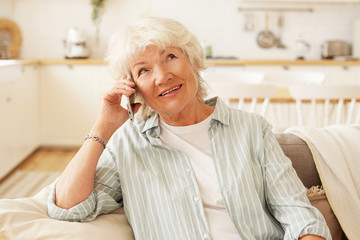 Wall Mural - Indoor shot of charming friendly senior gray haired woman holding generic smart phone close to her ear, having hearing problem, talking to her friend, sitting comfortably on couch in living room