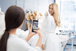 © pressmaster - Unrecognizable brunette taking photo of happy young woman trying on wedding gown in dress-making studio