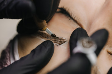 Hands in black gloves hold the manipulator with the microblading needle and draw her hair on the girl’s eyebrows close-up.