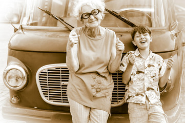 Wall Mural - Fashionable, cheerful grandmother with her grandchild are fooling around a retro car imitating driving. Summer family vacation and travel. Retro style. Sepia