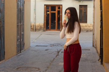 Portrait of attractive caucasian young woman model kissing euros banknotes, in the city, white top and red pants, dark air. Place for your text in copy space.