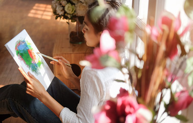 Cropped shot view of Woman painter sitting on the floor in front of the canvas and drawing. Artist studio interior. Drawing supplies, oil paints, artist brushes, canvas, frame.