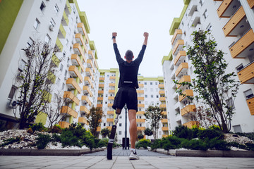 Wall Mural - Rear view of handsome sportsman with artificial leg standing with hands in the air outdoors surrounded by buildings.