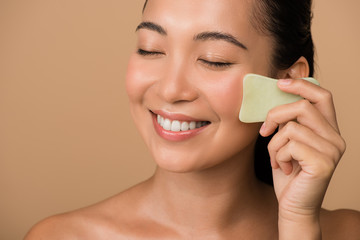 smiling beautiful naked asian girl with closed eyes using facial gua sha jade board isolated on beige