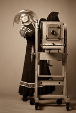 Vintage Retro Style Photo Of A Young Woman In A Long Modest Dress And Hat Standing Near A Daguerreotype Vintage Camera On A Gray Background. Retro Photography And Vintage Concept.