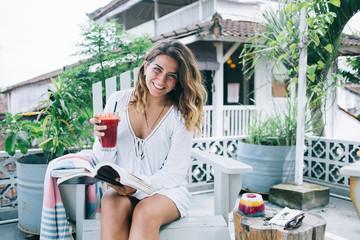 Positive adult lady reading book and drinking red smoothie on terrace