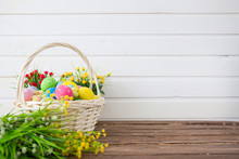 Colored Easter Eggs In The Basket And Spring Flowers On Wooden Background. Greeting Card