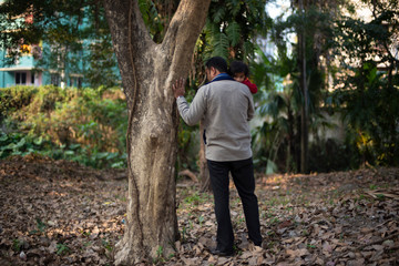 Back side of Indian brunette father taking his baby boy in lap in winter garments in winter afternoon on a dry grass field in green forest background. Indian lifestyle and parenthood.