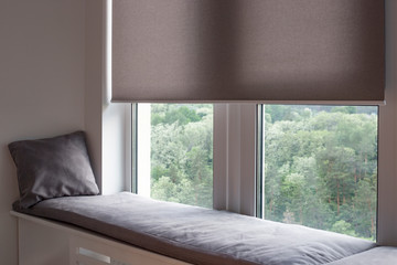 Poster - Motorized roller shades. Automatic roller blinds beige color on large windows. Remote Control Shades are near the sofa with the pillow. Sofa covered with pastel velours. Summer. Green trees outside.