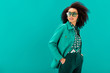 © LIGHTFIELD STUDIOS - stylish african american woman in jacket with hands in pocket looking away isolated on turquoise