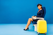 © LIGHTFIELD STUDIOS - african american flight attendant sitting on seat near travel bag on blue background