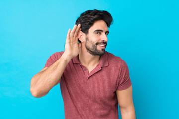 Wall Mural - Young handsome man with beard over isolated blue background listening to something by putting hand on the ear