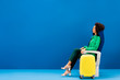 © LIGHTFIELD STUDIOS - smiling african american woman sitting on seat near travel bag on blue background