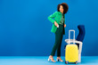 © LIGHTFIELD STUDIOS - smiling african american woman putting in pocket smartphone and standing near travel bag and seats on blue background