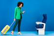 © LIGHTFIELD STUDIOS - smiling african american woman holding travel bag and using smartphone on blue background