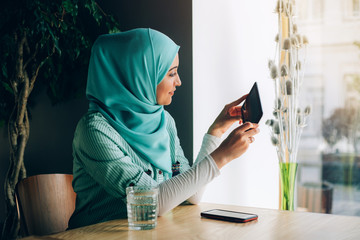 Wall Mural - Beautiful young arabic girl in hijab posing for camera at cafe with digital tablet near the window.