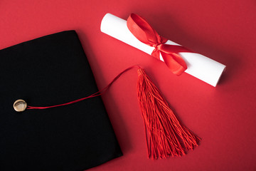 Wall Mural - Top view of diploma with beautiful bow and black graduation cap with tassel on red background