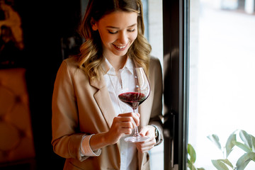 Single pretty young woman with glass of red wine standing near window and looking aside