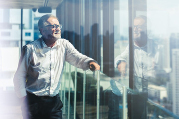 Retirement concept. Senior grey-haired businessman standing and looking to right hand at modern business lounge high up in an office tower.