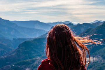 A close-up back view of a young redhead woman with her long hair fluttering in front of the mountain valley landscape from behind on a sunny windy day