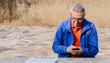 © Cavan Images - Senior man looking and touching screen of smartphone while sitting on park