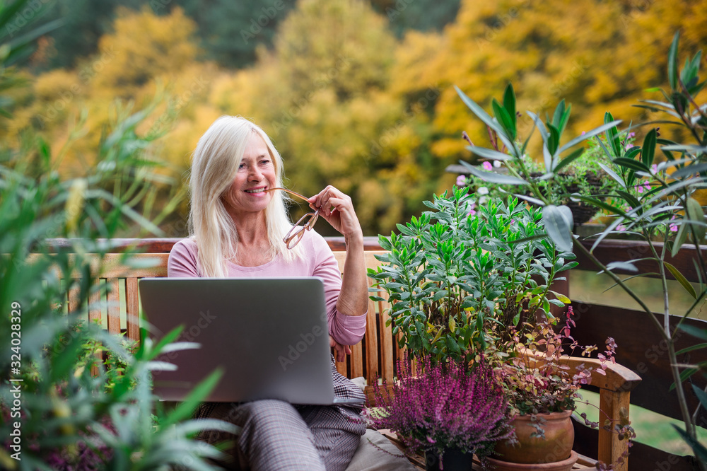 Senior woman with laptop sitting outdoors on terrace, working. - obrazy, fototapety, plakaty 