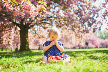 Cute one year old girl sitting on the grass and eating strawberries