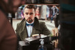 © Fxquadro - Stylish and elegant young male customer sitting outdoors next to coffee making barista in a mobile coffee shop in a city emporium, wearing green wool coat, white shirt and bow tie, holding a paper cup